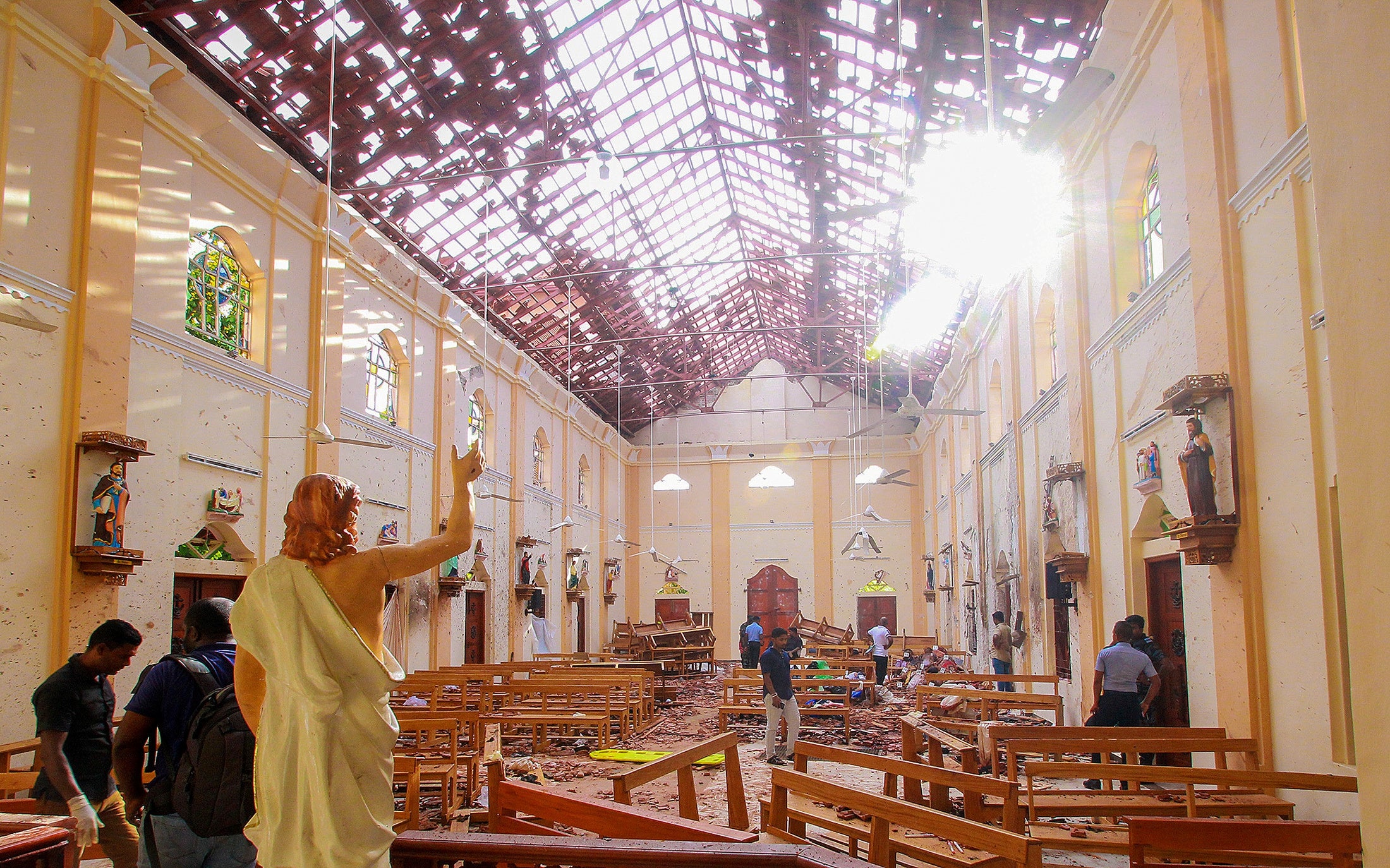 A blood-spattered statue of Jesus Christ while crime scene officials inspect the site of a bomb blast, as the sun shines through the blown-out roof, inside St Sebastian's Church in Negombo