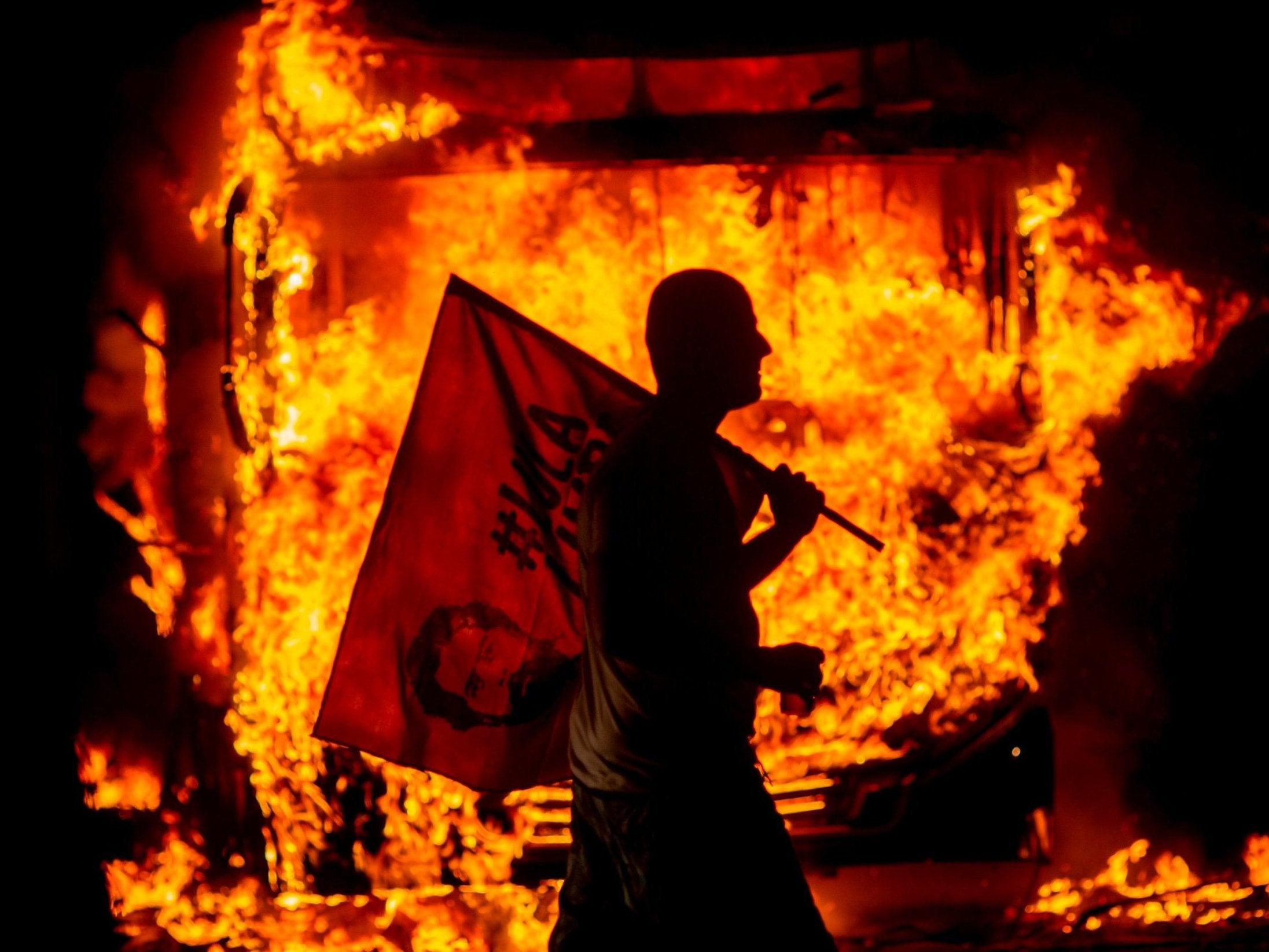 A man holds a flag and walks past a bus in flames during a protest