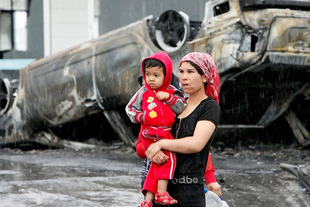 An Uighur woman and child walk past a burned car following riots in Urumqi 10 years ago