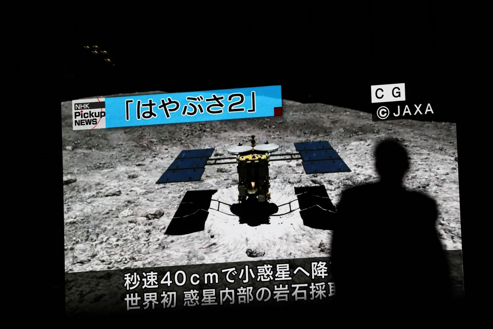 In this picture taken on July 10, 2019 a man walks past a screen displaying a computer-generated image of the Hayabusa2 probe, during a news broadcast at Akihabara district in Tokyo