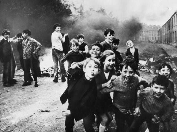 A group of children playing up to the camera while a fire smoulders in the street in Belfast, 1971