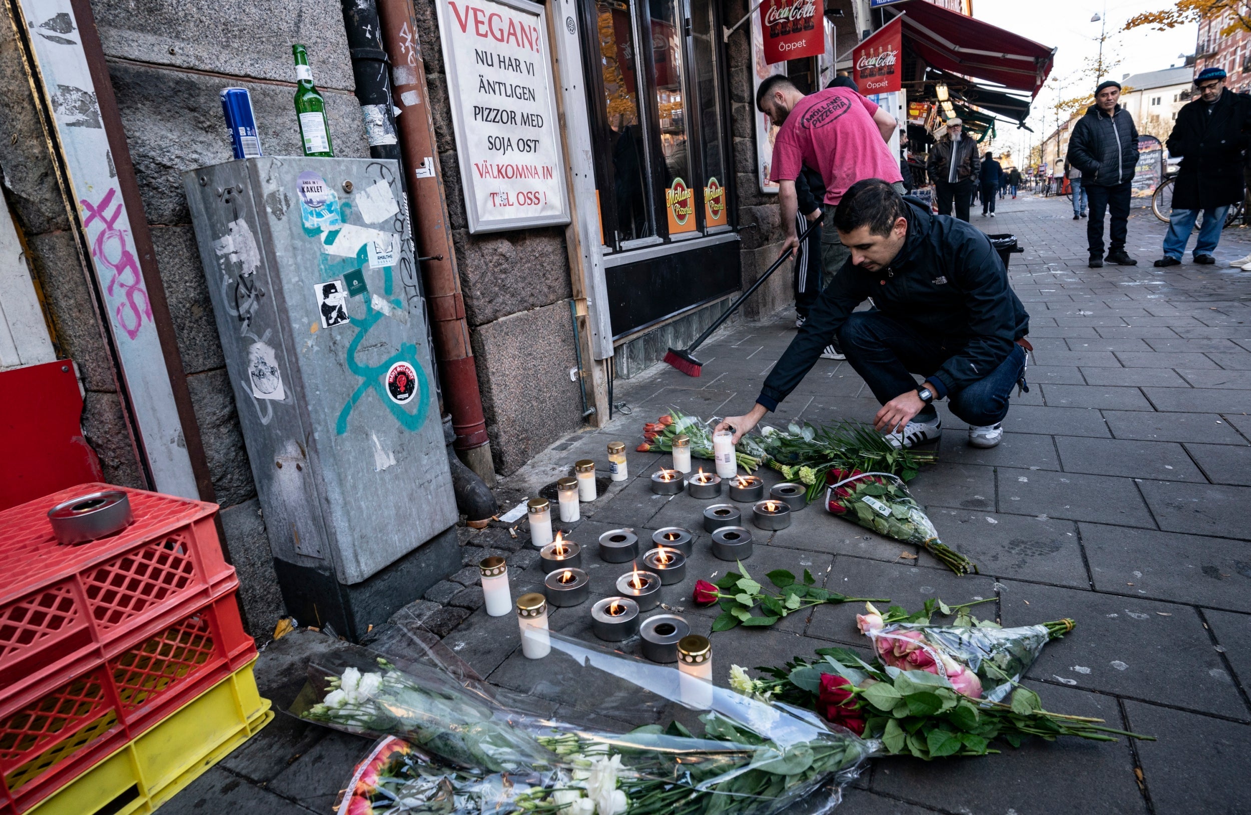 A man places flowers in tribute to victims of a shooting while a restaurant employee sweeps broken glass at the crime scene in Malmo, Sweden