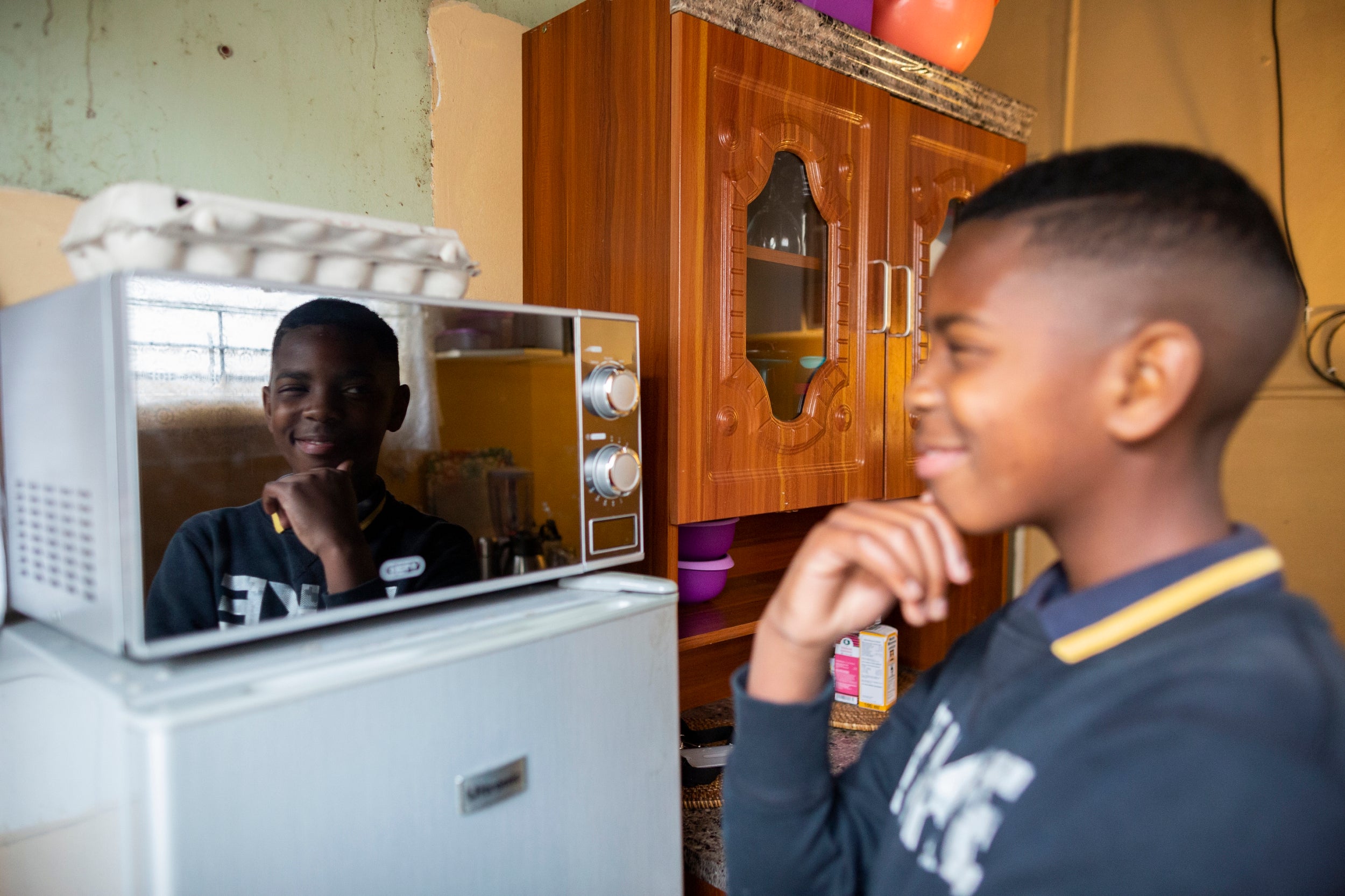 A boy living on the Autism spectrum interacts with his reflection in a microwave oven in the kitchen of his home in Khayelitsha
