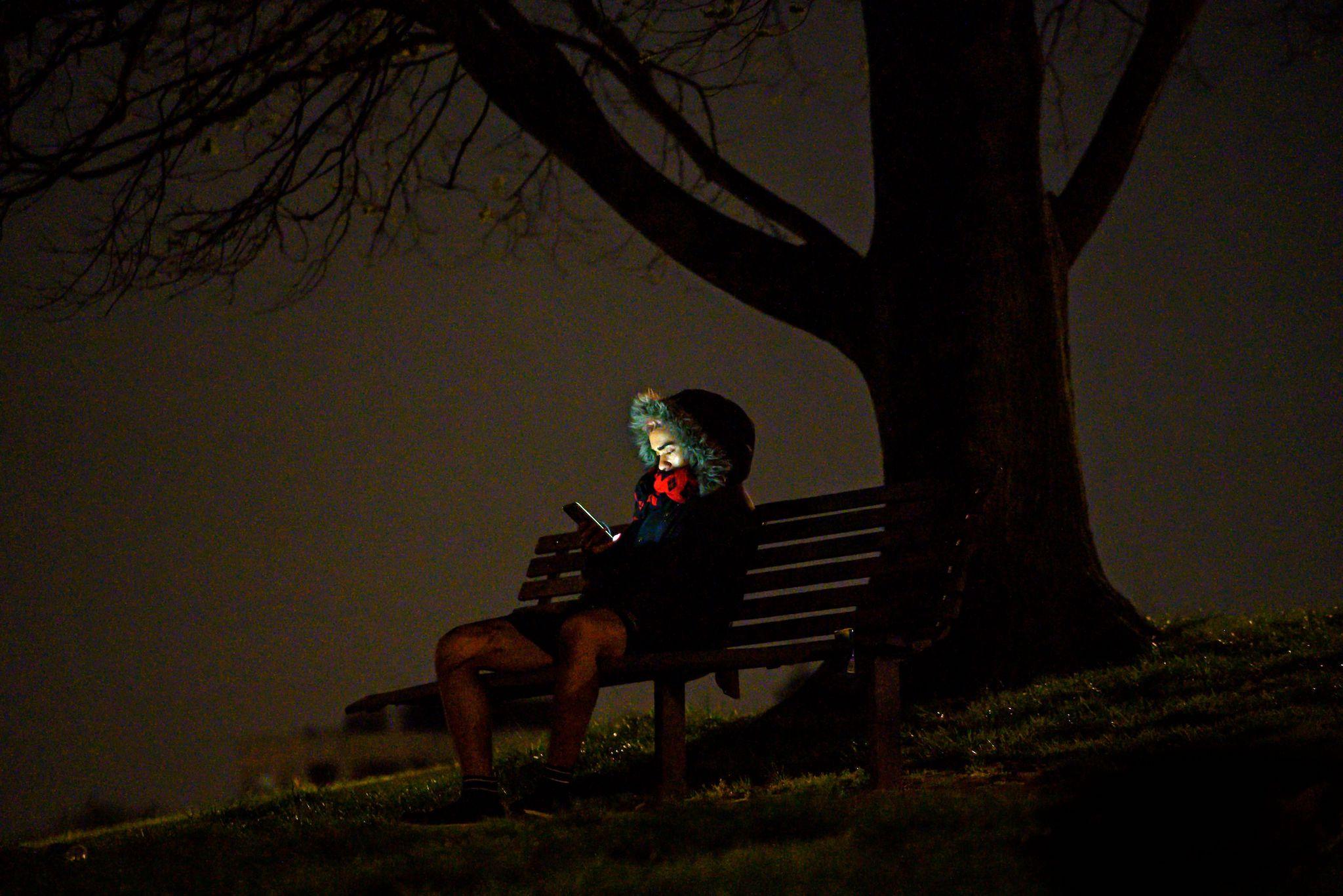 A man sits on a bench looking at his mobile phone at night on Primrose Hill in London on April 18, 2020, during the novel coronavirus COVID-19 pandemic
