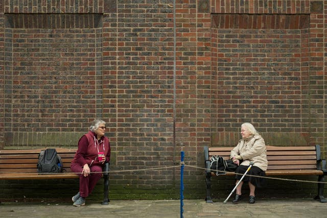 Two women observe social distancing measures as they speak to each other from adjacent park benches