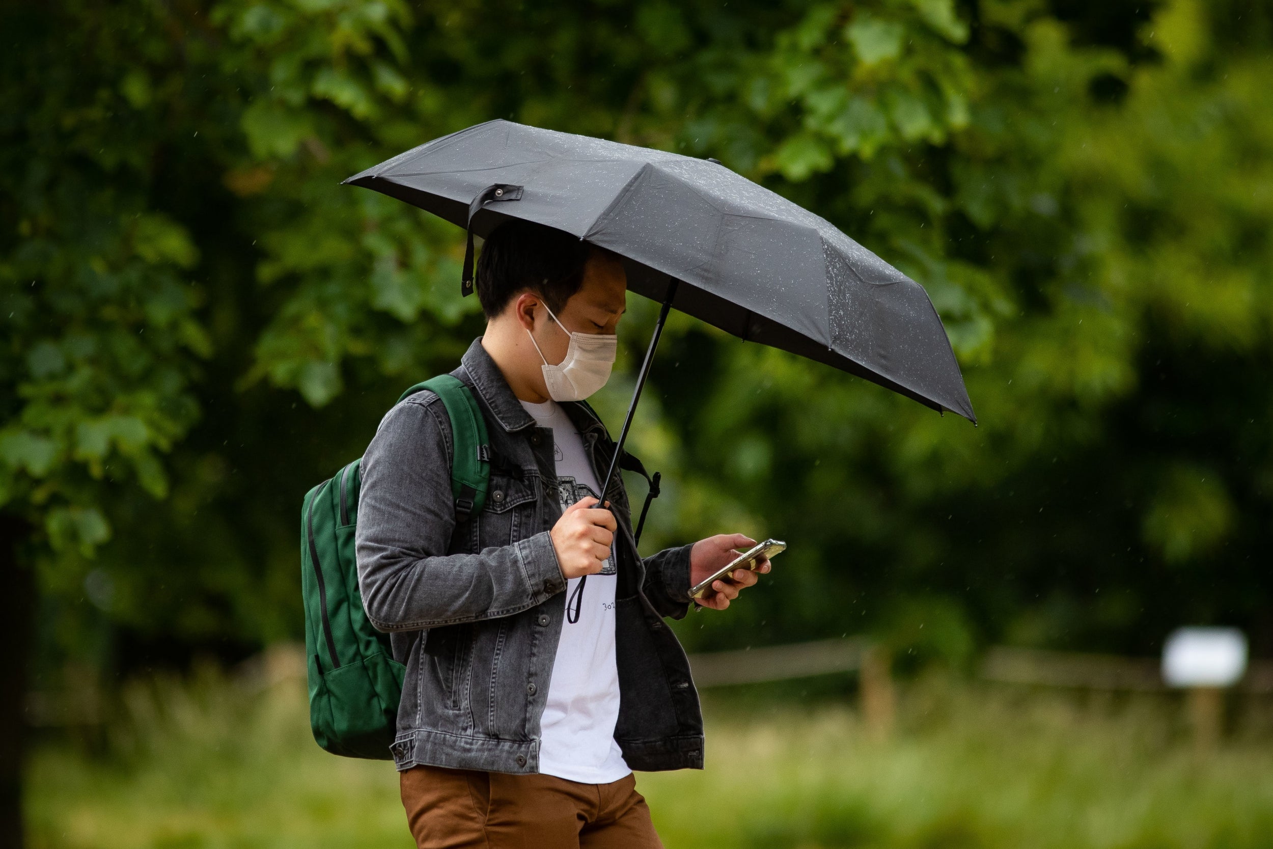 A man wearing a face mask shelters from the rain under an umbrella in London