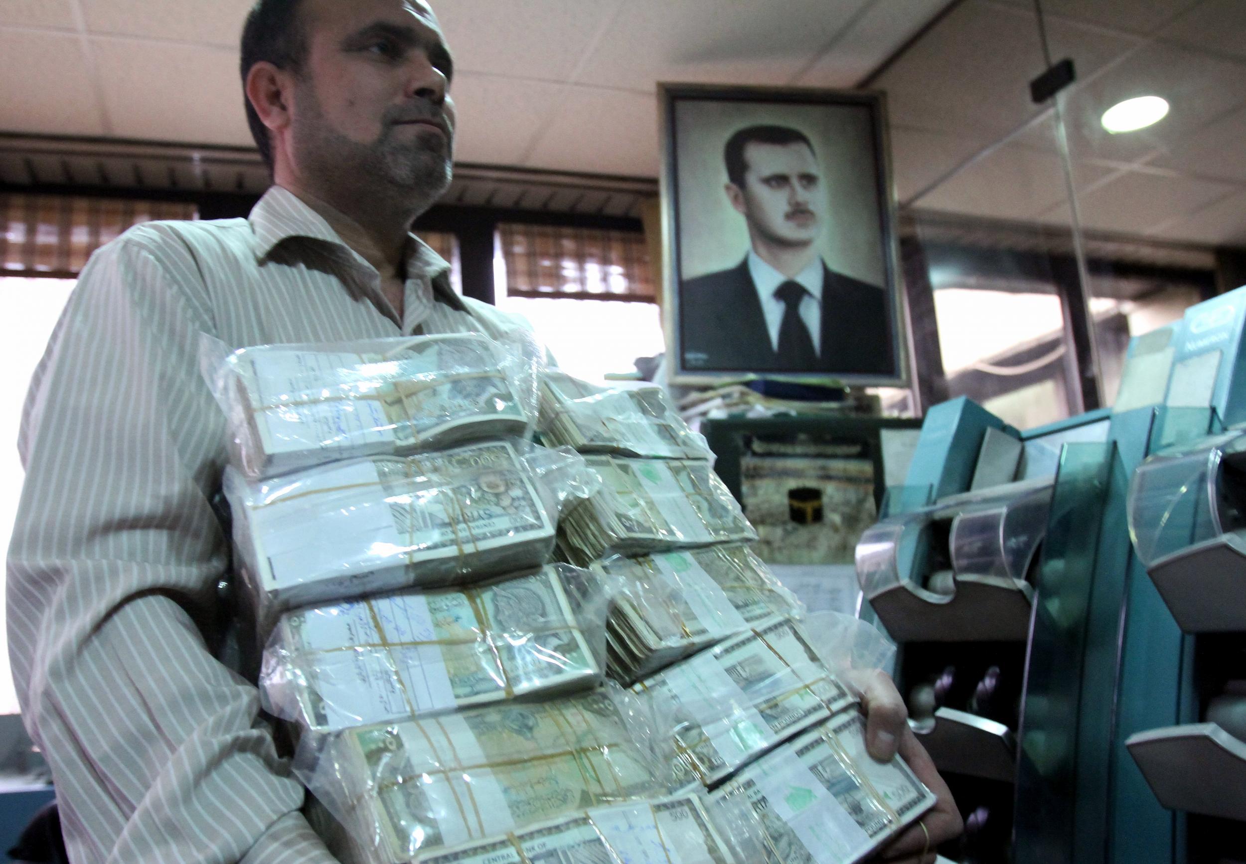 A banker carries a load of money as he walks past an image of the Bashar al-Assad at a bank in the capital in Damascus (Anwar Amro/AFP)