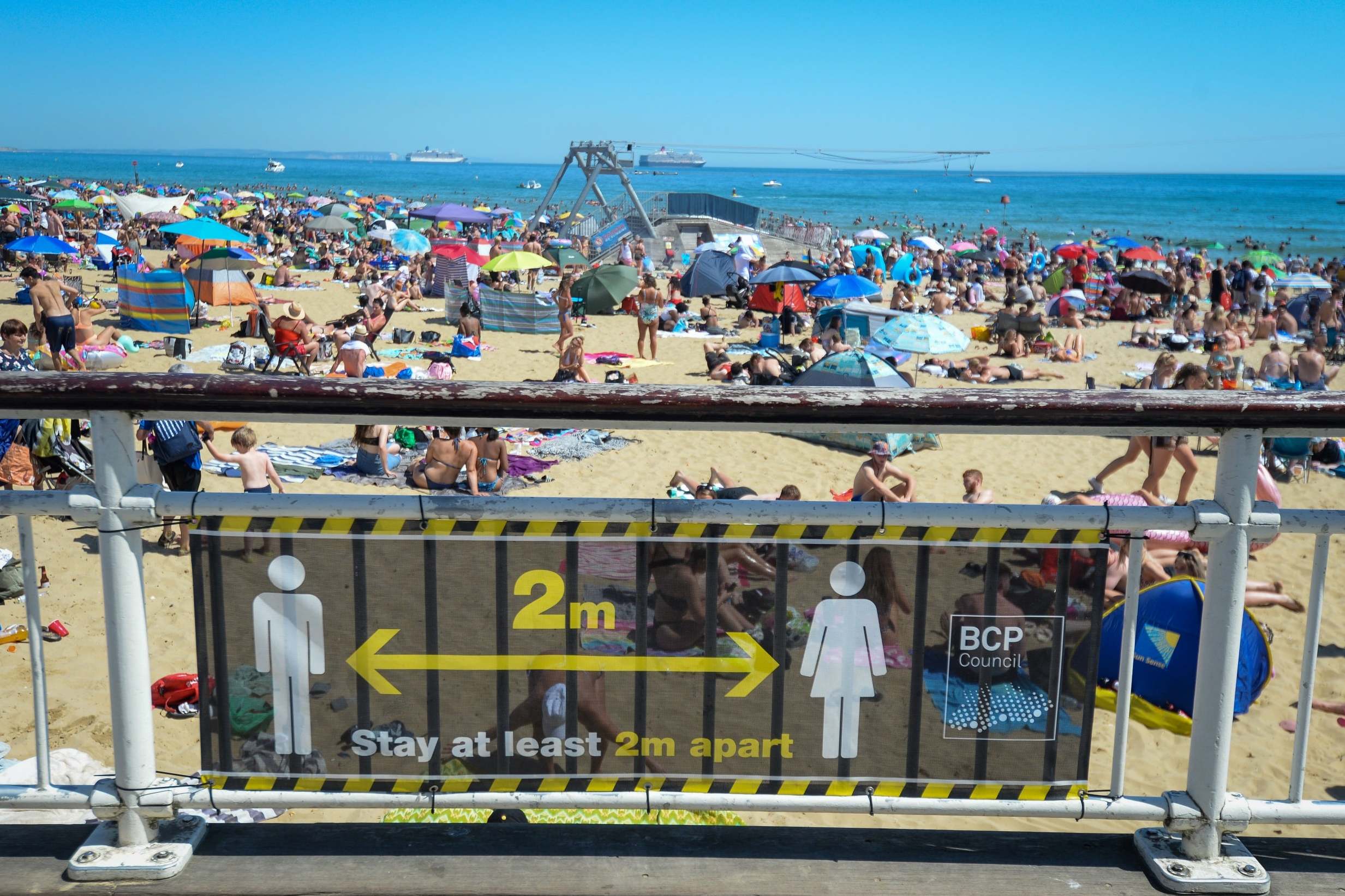 A social distancing warning sign is displayed against a backdrop of a crowded Bournemouth beach